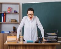young-woman-teacher-wearing-glasses-explaining-lesson-angry-frustrated-going-mad-sitting-school-desk-front-blackboard-classroom