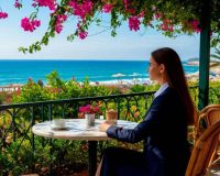 Девушка в деловом костюме любуется океаном, сидя на веранде летнего кафе